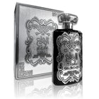 The image displays a product set consisting of a perfume bottle and its packaging box. Both the bottle and the box are adorned with elaborate embossed silver designs. The central motif is a stylized, ornate emblem with what appears to be Arabic script at the center, possibly the brand or fragrance name, 'AL IBDA'.  The box has a textured surface with the same motif repeated in a more subdued pattern in the background. 