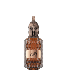 A product shot of Le Falcone Niche Collection’s “Al Majd” perfume bottle against a white background: a tall, amber-colored glass vessel with a diamond-cut texture and a polished bronze plaque displaying Arabic calligraphy above the words “Al Majd.” It’s crowned by an ornate bronze cap sculpted into a stylized warrior’s helmet with a crest-style loop at the top, conveying a sense of luxury and strength.
