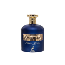 The bottle is a deep cobalt blue with a smooth, rounded shape. It has a metallic gold label with the name "AMBERLEY" embossed in the center. Below the main label, in elegant script, is the name "Ombre Blue." A small, gold stag emblem is placed at the bottom center of the bottle, indicating the logo for "MAISON ALHAMBRA." The cap of the bottle is gold with a textured design, resembling a crown or cog. 