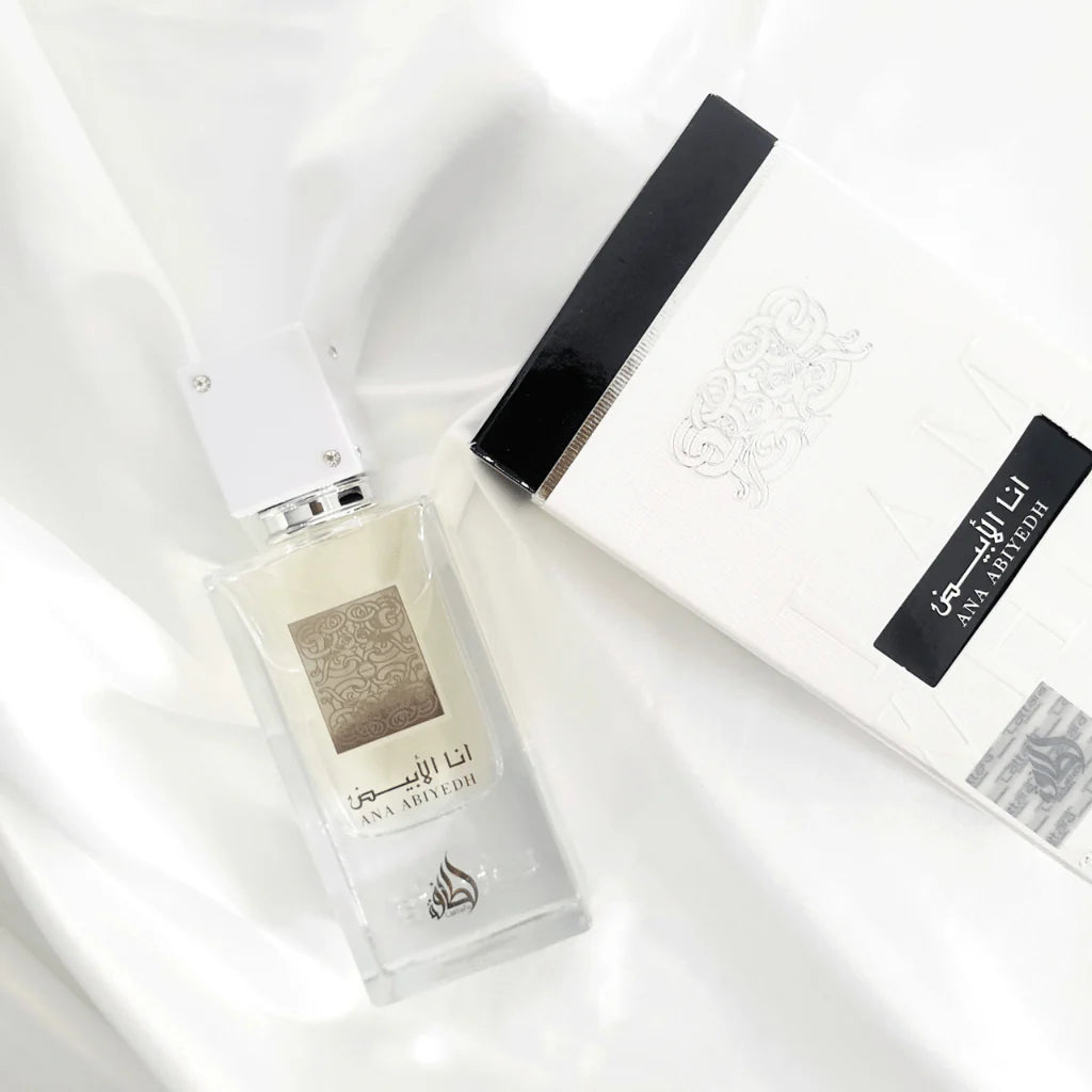 A clear rectangular bottle of Ana Abiyedh Eau de Parfum by Ard Al Zaafaran Lattafa Perfumes, lying next to its elegant white box with black accents. The bottle features a minimalist design with a white cap adorned with small silver accents. The front of the bottle displays intricate silver patterns along with the fragrance name "Ana Abiyedh" in both Arabic and English. The packaging box mirrors the bottle's sophisticated design with embossed details and the brand name.