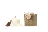 The image shows a luxurious product presentation featuring a candle and its packaging. On the left, there is a cream-colored, multi-tiered candle topped with a golden accent and adorned with a dark brown tassel. The candle sits on a reflective surface that enhances its elegance. On the right, there is a gold-colored box, which likely houses the candle, adorned with a beige ribbon and an attached card with additional branding details.