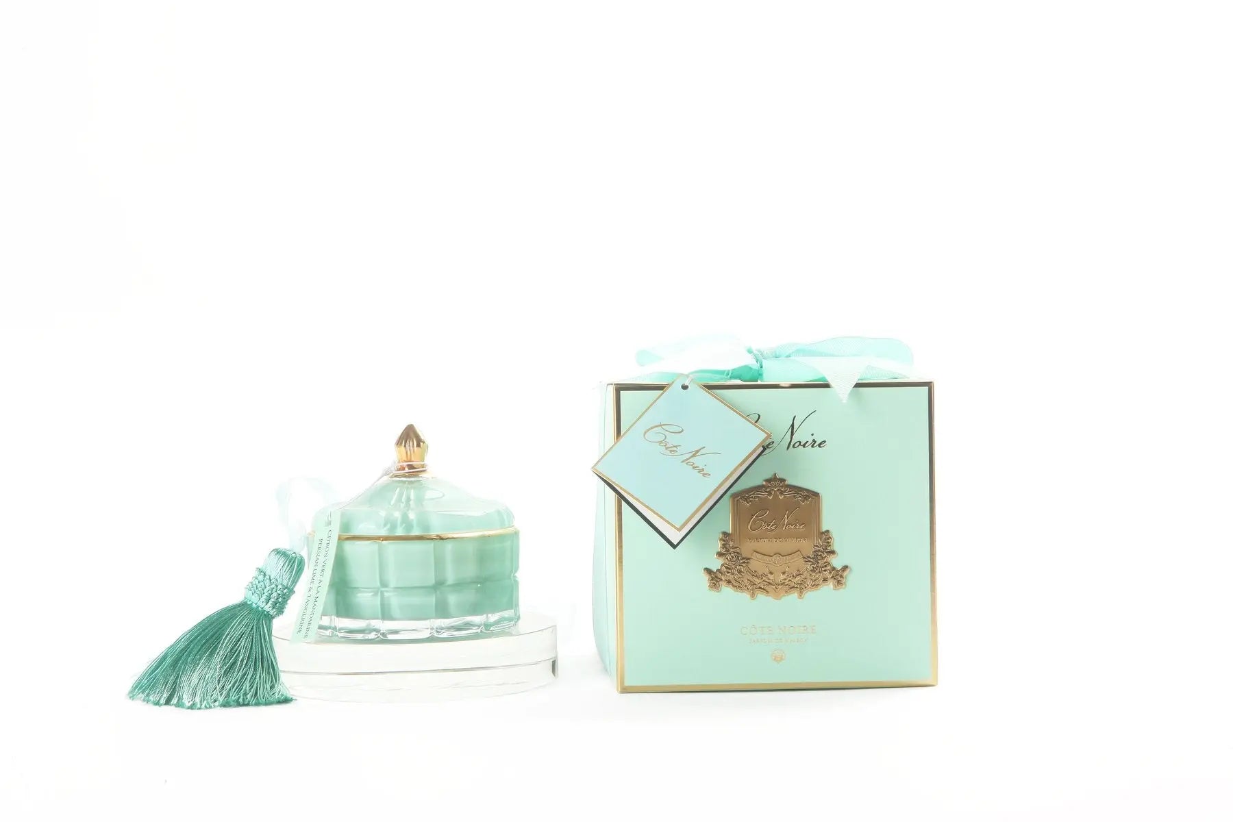 The image features a luxurious candle and gift box display. On the left, there's a decorative candle in a translucent Tiffany blue glass container adorned with a golden tassel. The lid of the candle has a golden knob on top, adding a touch of elegance. On the right, a matching Tiffany blue gift box with a golden crest and a white ribbon is shown. The box also has a gift tag attached with a light blue ribbon, inscribed with "Cote Noire."