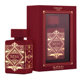 This image shows the "BADEE AL OUD SUBLIME" perfume by Lattafa, placed next to its packaging. The perfume bottle is deep burgundy with a matte finish and a square cap, featuring gold geometric and traditional designs with the name in Arabic and English. The packaging matches the bottle's color and design, with a rectangular box that has a window display framed by intricate gold patterns and the name "SUBLIME" printed in gold at the top.