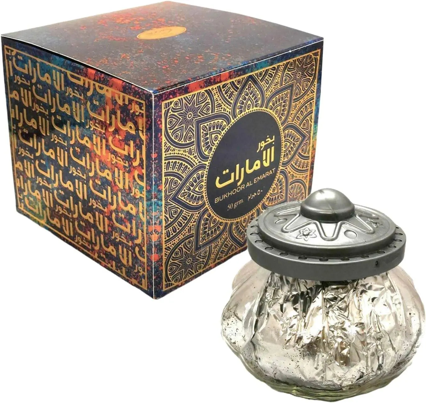 The image features "Bukhoor Al Emarat" by "Ard Al Zaafaran", which is presented as a luxurious traditional fragrance product. There is a cubic box with an elaborate pattern in gold, blue, and orange hues, featuring Arabic calligraphy and English text on its side, which indicates the product name. The box is open, revealing a rich, textured black interior. Accompanying the box is a decorative glass burner with a silver foil texture and a dark, metallic lid with ornamental designs.