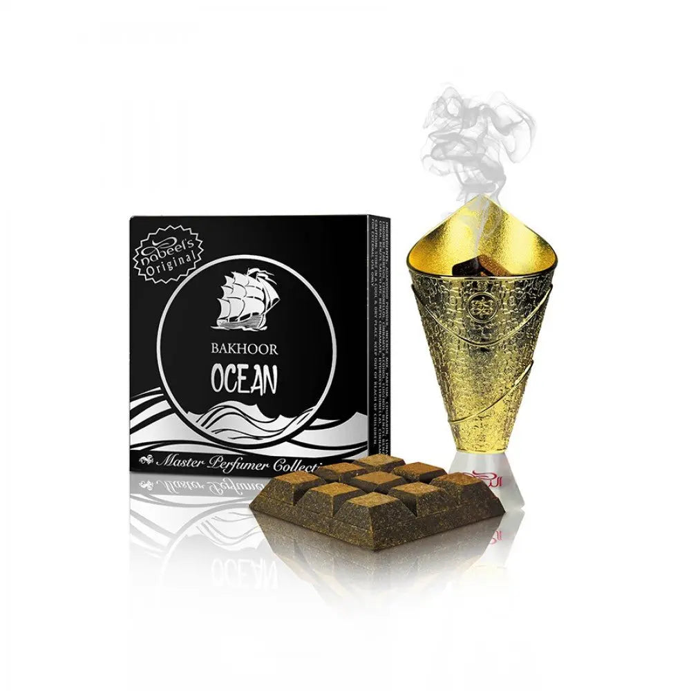 The image features "Bakhoor Ocean" from Nabeel Perfumes' Master Perfumer Collection. On the left is a black box with a stylized white graphic of a sailing ship and waves, encapsulated within a round label that reads "Bakhoor Ocean". To the right, there's a golden bakhoor burner with an ornate design, from which smoke is gracefully curling upward, suggesting the bakhoor is in use. In front of the box lies a collection of brown bakhoor bricks, arranged in a geometric pattern.