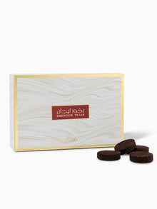 The image presents a box of Bakhoor Tejan by Rasasi Perfumes. The box is rectangular with an elegant white and grey wood grain effect and a sophisticated golden border. The product's name, "Bakhoor Tejan," is printed in the center in a rich burgundy rectangle with both Arabic and English lettering. At the bottom of the box is a subtle, decorative golden motif. In front of the box, there are multiple dark brown, circular bakhoor incense tablets.