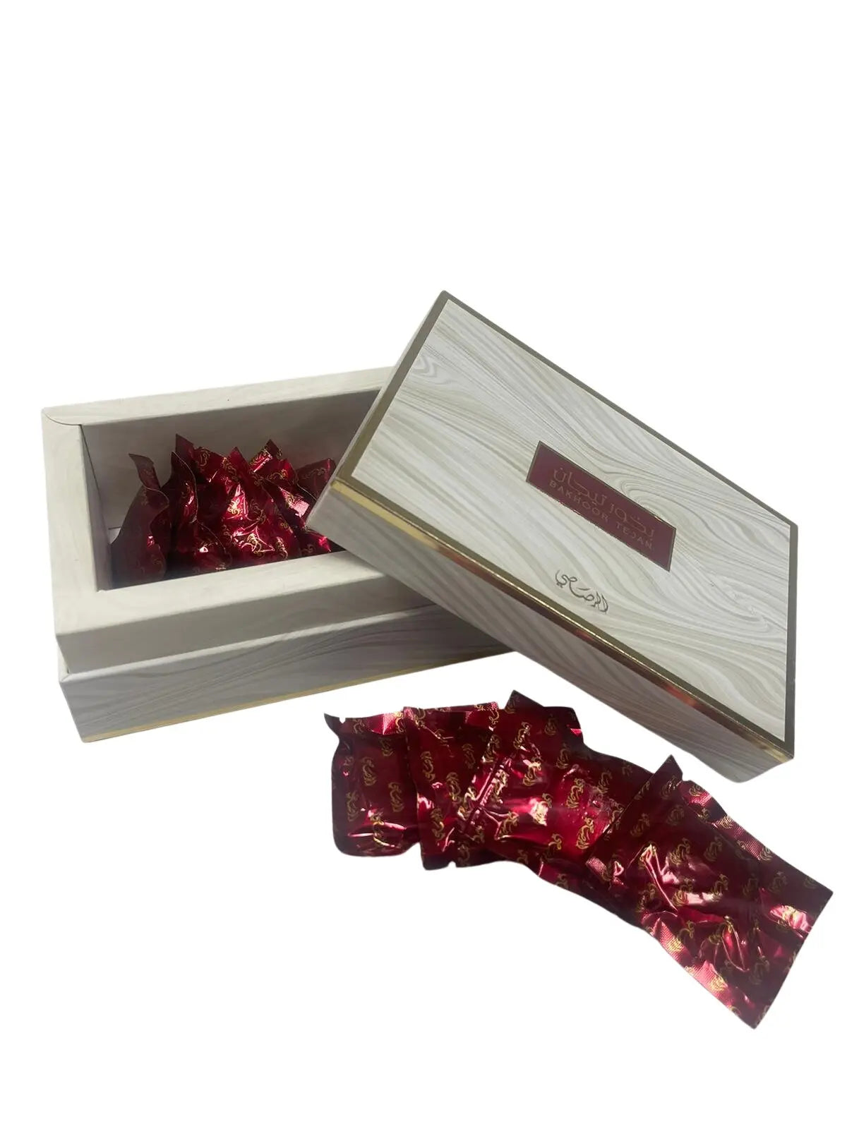 A wooden box with a slide-out lid partially opened, revealing multiple red foil-wrapped items with gold print, likely to be bakhoor or scented bricks used for fragrance. The lid of the box has Arabic script and English text which may indicate the brand or type of the product, suggesting it's a commercial product packaging.