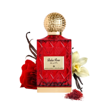 The image depicts a red glass perfume bottle with a golden, textured cap, labeled "Balas Rose" in English with Arabic script above it, suggesting the fragrance's name by the brand Ibraheem Al Qurashi.   In the composition, there's a red rose to the left, suggesting the scent's floral nature, vanilla beans to the left, indicating a warm, sweet note, a white flower (possibly vanilla or orchid) to the right, and a small pile of red saffron threads in the front right, hinting at the luxurious ingredients.