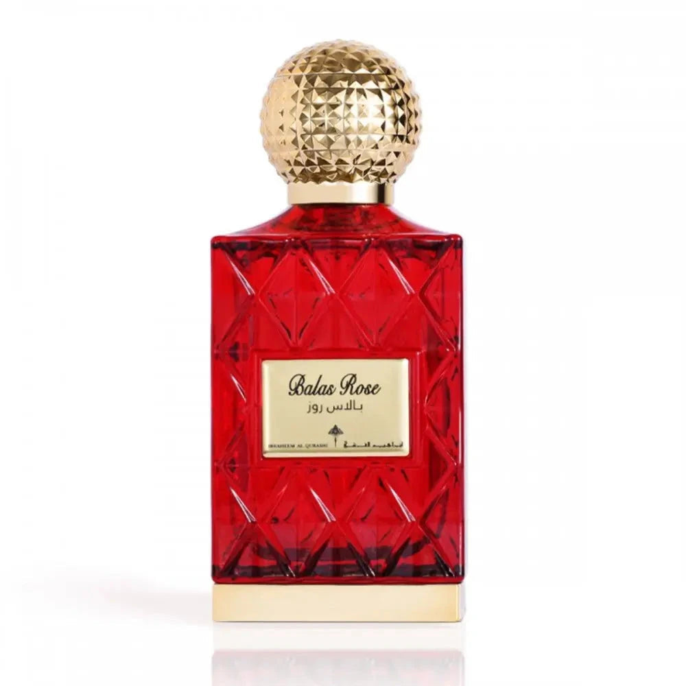 The image shows a red glass perfume bottle with a golden, textured cap. The label on the bottle reads "Balas Rose" in English with Arabic script above it, indicating the fragrance name. The bottle has a faceted design that gives it the look of a cut gemstone. The bottle is placed against a white background, which contrasts with the red color of the glass, highlighting the bottle's luxurious appearance. The golden cap and label add an elegant touch to the overall presentation of the perfume.