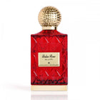 The image shows a red glass perfume bottle with a golden, textured cap. The label on the bottle reads "Balas Rose" in English with Arabic script above it, indicating the fragrance name. The bottle has a faceted design that gives it the look of a cut gemstone. The bottle is placed against a white background, which contrasts with the red color of the glass, highlighting the bottle's luxurious appearance. The golden cap and label add an elegant touch to the overall presentation of the perfume.