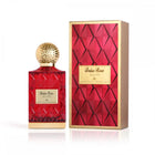 The image features a red glass perfume bottle with a golden, textured cap, alongside its matching packaging. The label on the bottle has "Balas Rose" written in English and Arabic, signifying the fragrance's name by the brand Ibraheem Al Qurashi. The design of the bottle features a faceted pattern, giving it a luxurious, gem-like appearance. The packaging box is a complementary gold color with a red faceted design and a label that matches the bottle's. 