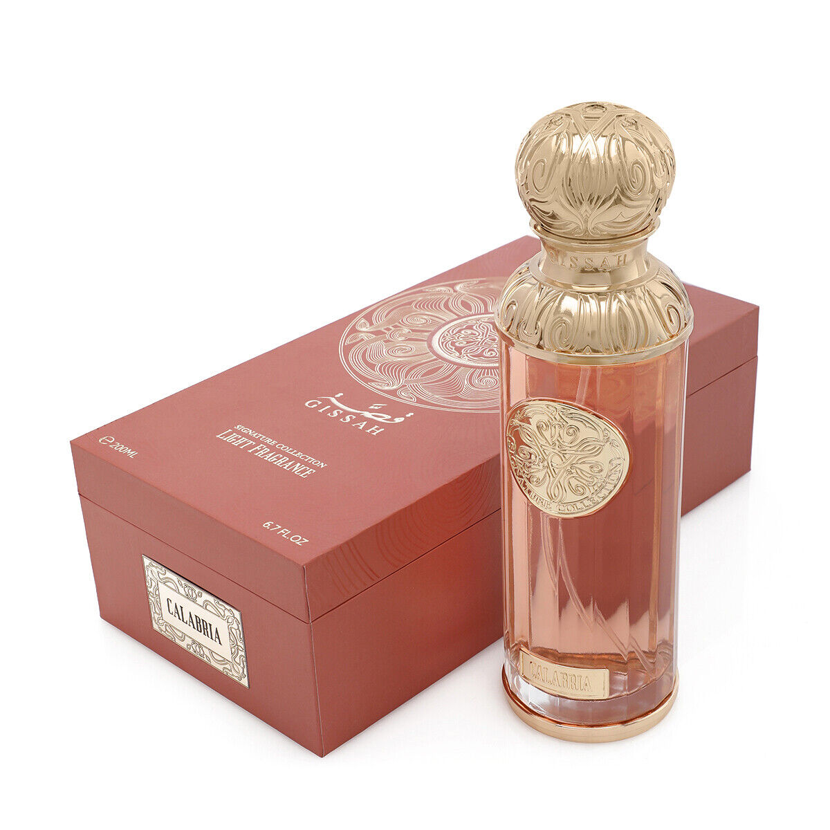 The image shows a perfume bottle named "Calabria" from the Gissah Signature Collection. The bottle is tall and cylindrical with a clear glass body that has a slight rose tint, and it is adorned with a gold ornate medallion that features intricate designs. The cap is a luxurious gold color with a decorative embossed pattern. Beside the bottle is the packaging, a box in a matching rose tint with a gold mandala design and the names "Gissah" and "Calabria" printed on it.