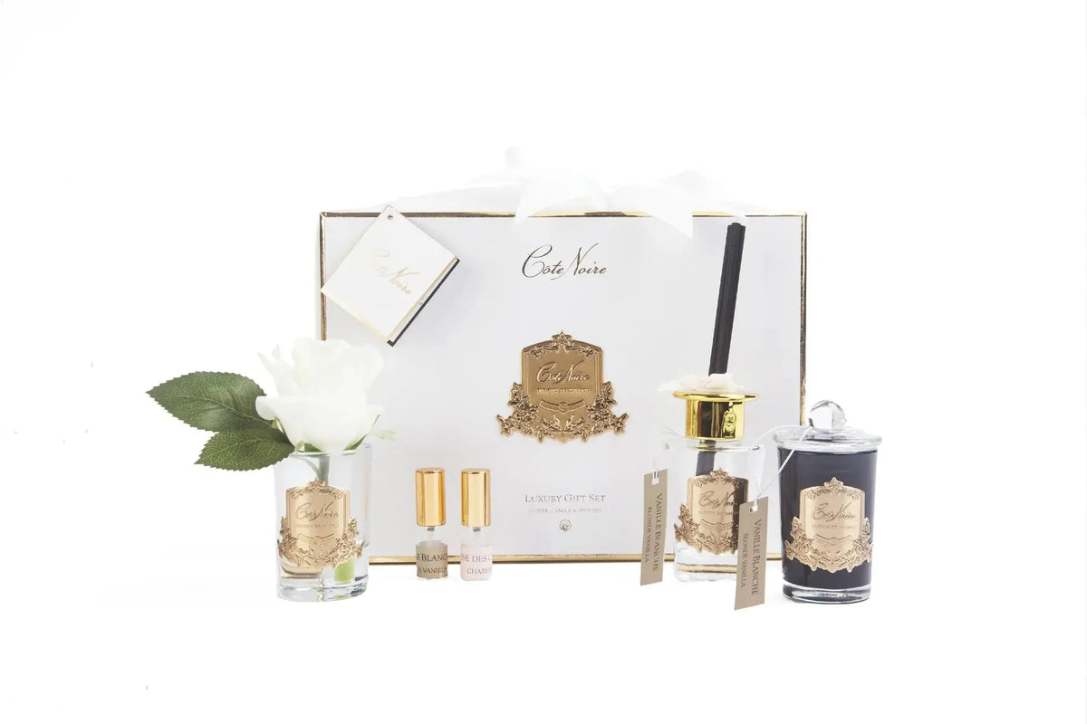 The image displays an elegant and sophisticated luxury gift set from Cote Noire. In the foreground, a clear glass vase with a single white rose complements the set. The main feature is a large white gift box with gold edges and a central gold crest, labeled "Cote Noire." Accompanying the box are several perfume products, including two small gold-capped perfume bottles, a black glass candle with a clear lid, and a diffuser with a black reed.
