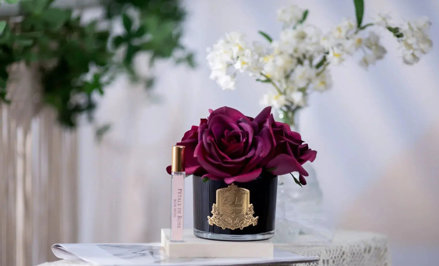 The image features a sophisticated product presentation on a marble table, bathed in soft, natural light. In the foreground, a black glass vase with a golden crest holds a large, lush deep red rose. Beside the vase is a sleek perfume roller with a clear body and a golden cap, labeled "Petale De Rose." In the background, a small vase filled with white flowers adds a delicate touch to the composition.