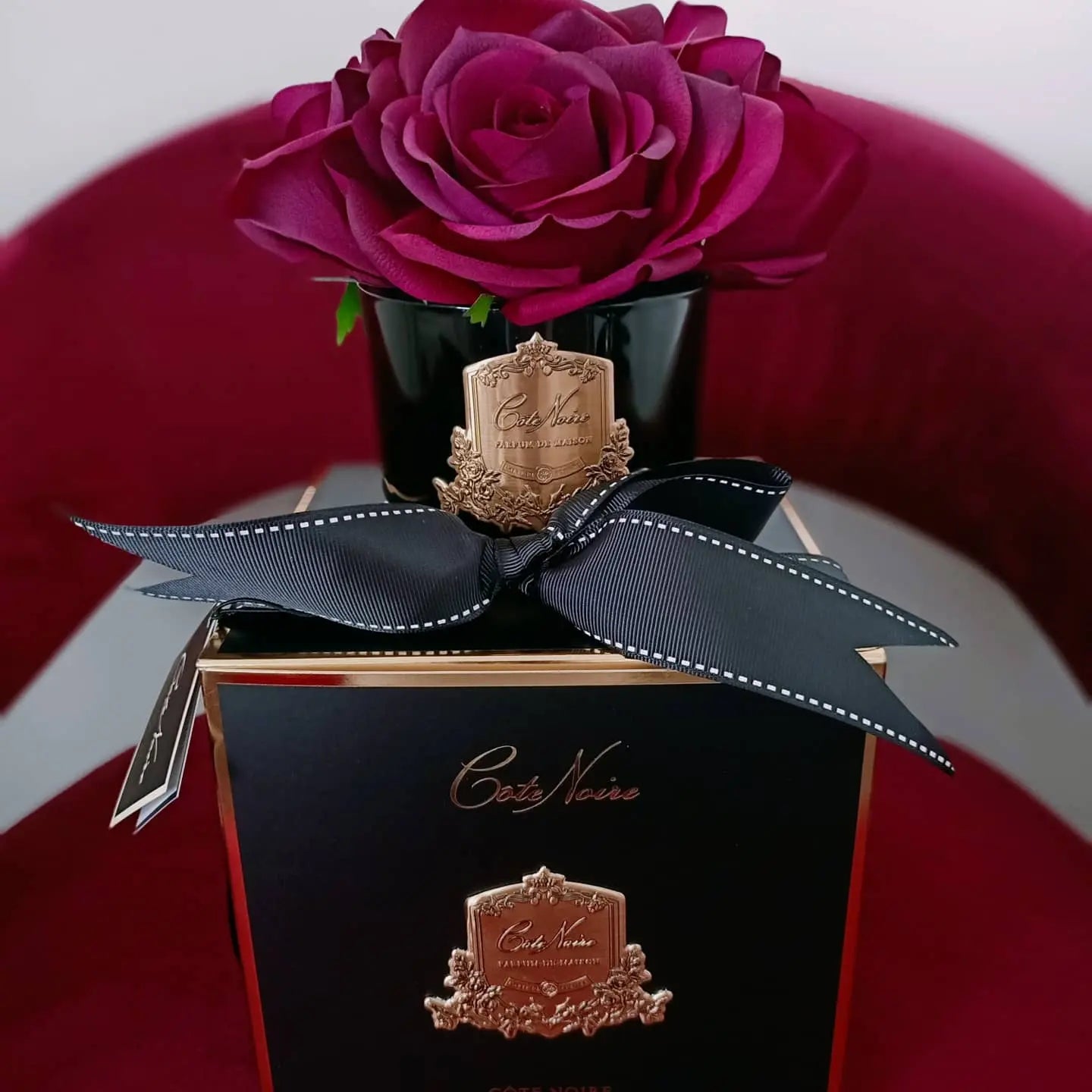 The image showcases an exquisite product display against a rich, red velvet background. A black glass vase holds an intricately detailed deep purple rose, sitting atop a black and gold gift box. The box is elegantly tied with a black ribbon featuring a white edge and adorned with a gift tag inscribed with "Cote Noire." The gold crest on both the vase and the box adds a touch of opulence to the overall presentation, emphasizing luxury and sophistication.