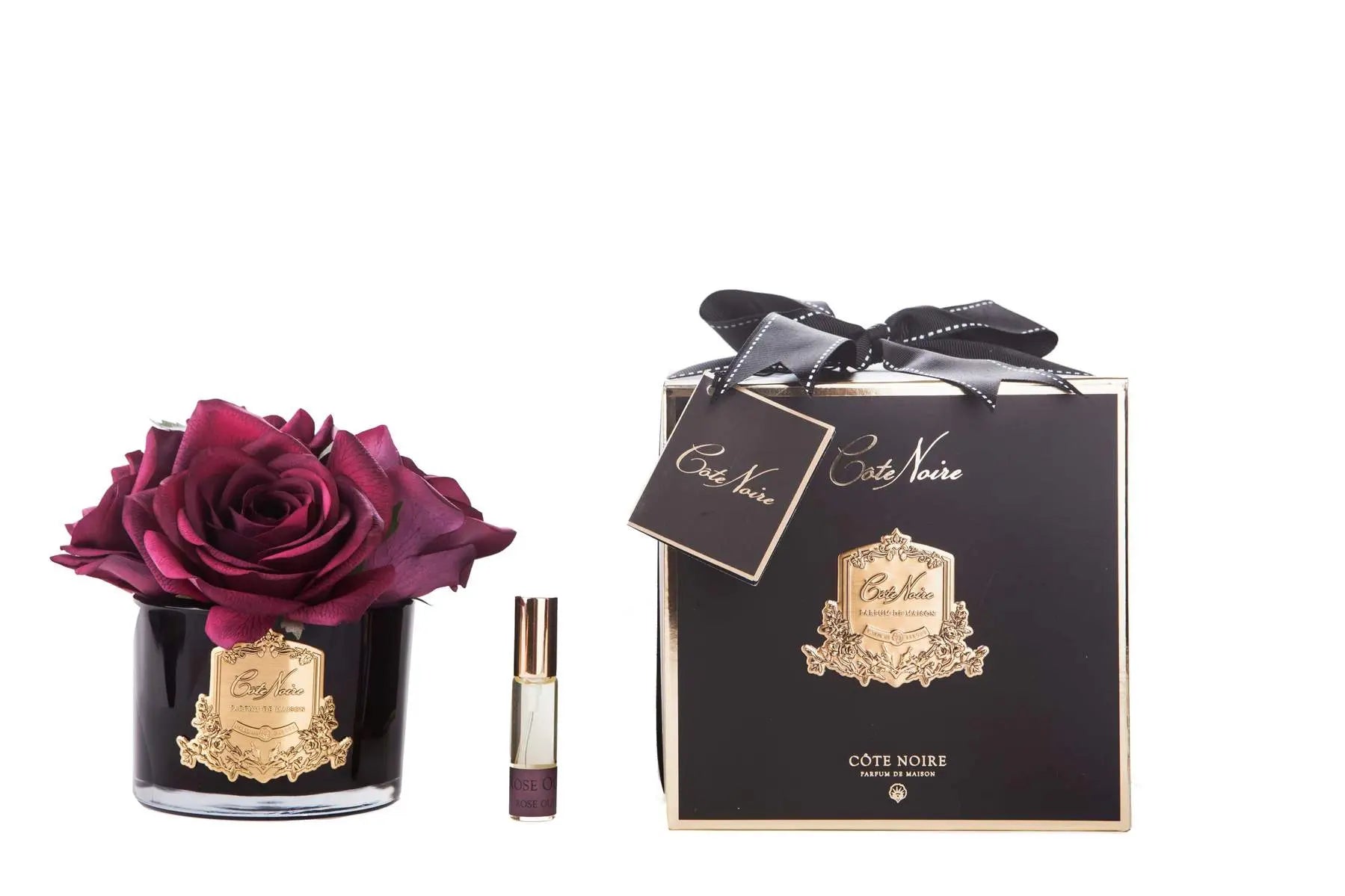 The image presents an elegant product arrangement against a white background. On the left, a black glass vase contains a single large deep red rose, beautifully detailed, with a gold label reading "Cote Noire Parfum de Maison." Beside the vase is a small, stylish perfume roller with a clear body and a gold cap, labeled similarly. On the right, a luxurious black gift box with a golden crest design is tied with a black ribbon and adorned with a gift tag reading "Cote Noire."