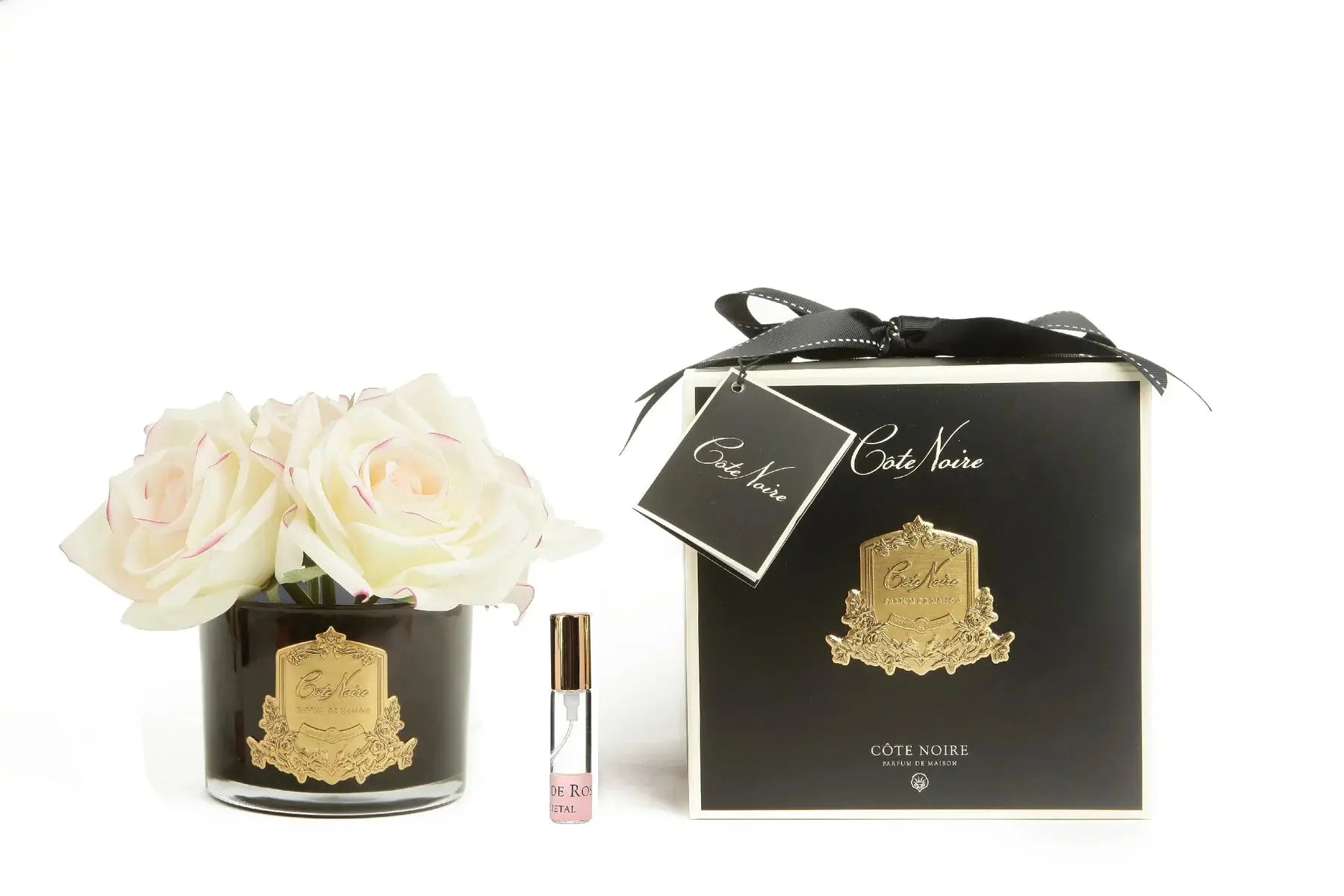 The image features a luxurious product arrangement against a white background. On the left, there is a black glass vase filled with several light pink roses, the vase adorned with a gold label reading "Cote Noire Bougie de Jardinia." Beside the vase, there is a small, elegant perfume roller with a clear body and a gold cap, labelled similarly. On the right side of the image, there's a sleek black gift box with a golden crest design, tied with a black ribbon and a gift tag that reads "Cote Noire."