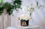 The image captures a delicate and sophisticated product display on a marble table. In the foreground, there is a black glass vase adorned with a golden crest, holding several light pink roses. Beside the vase is a sleek perfume roller with a clear body and a golden cap, labeled "Petale De Rose." In the background, a blurred arrangement of white flowers adds a soft, serene atmosphere to the scene.