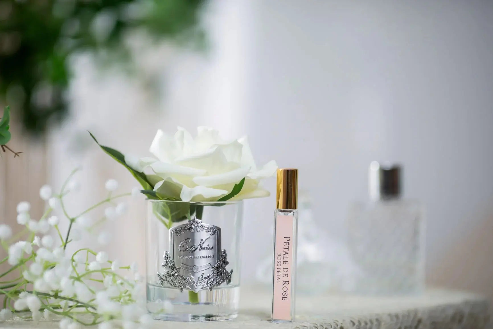 A Cote Noire product featuring a single white artificial rose in a clear glass jar with a silver label, alongside a small vial of perfume labeled 'Pétale de Rose' with a gold cap. The items are elegantly displayed on a lace surface, with a blurred background of greenery and additional perfume bottles, creating a delicate and luxurious setting.
