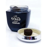 A navy blue jar with a silver lid open to reveal its contents, labeled "Bukhoor Dalua" in white Arabic and English script. In front of the jar sits a silver lid with a piece of bakhoor on it, a compressed incense cube used for scenting spaces. The design suggests a contemporary product grounded in traditional Middle Eastern fragrance practices.