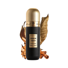 A cylindrical black perfume bottle with a gold cap, labeled "DOMINICAN TOBACCO" in white text, and "Extrait de Parfum 100 ml 3.4 fl.oz" in smaller white text. It has an Arabic inscription and the brand name "IBRAHEEM AL QURASHI" at the top. The bottle is presented in front of two large dried tobacco leaves and rests on a surface with scattered cinnamon sticks. The background is white, emphasizing the product.