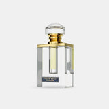 The image displays a modern and minimalist perfume bottle. The clear glass bottle has a square base and a slightly narrower top, with a central transparent chamber that holds the yellowish perfume liquid. It features a thick, gold band around the neck and is topped with a large, circular, transparent cap that resembles a cut gemstone.  The design of the bottle is clean and geometric, with a plain white background that accentuates the bottle's transparency and the liquid's color.
