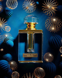 The image features an elegant perfume bottle prominently displayed against a dark blue background adorned with golden wireframe spheres that resemble festive ornaments. The bottle has a clear, rectangular shape with a thick base, enhancing its luxurious appearance. The cap is a large, transparent, faceted piece that resembles a cut gemstone, and it sits atop a golden neck, creating a striking contrast with the deep blue backdrop. 