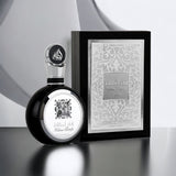 An image of a black and silver perfume bottle alongside its packaging. The bottle features a reflective black surface with a silver flower emblem and a silver cap with Arabic calligraphy. The box is a monochrome grey with elegant arabesque designs and the name "Fakhar Lattafa" in the center, along with "PRIDE OF LATTAFA" below it.