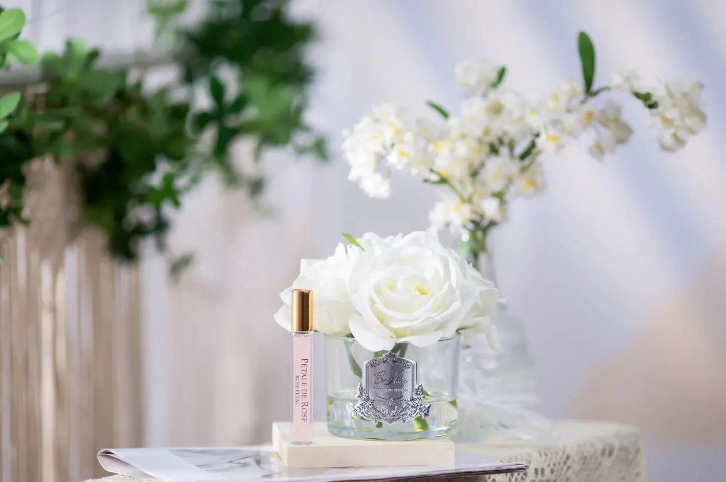 The image depicts a serene and graceful product presentation set on a marble table. In the foreground, a clear glass vase holds a large white rose, and beside it is a sleek perfume roller with a clear body and gold cap, labeled "Petale De Rose." In the background, a small glass vase with delicate white flowers adds to the floral theme.