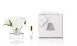 The image features a refined and elegant product display on a white background. To the left, there's a clear glass vase holding a single large white rose, with the vase labeled "Cote Noire Bougie de Jardinia." Next to it is a small, sleek perfume roller with a clear body and a gold cap, labeled similarly. On the right, there is a luxurious white gift box adorned with a white ribbon and a gift tag inscribed "Cote Noire."