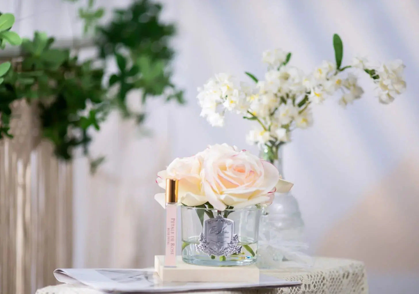 A Cote Noire product featuring five artificial pink blush roses in a clear glass vase with a silver crest, accompanied by a small vial of perfume labeled 'Pétale de Rose' with a gold cap. The items are displayed on a lace surface with a background of greenery and a vase of white flowers, creating a delicate and elegant setting.
