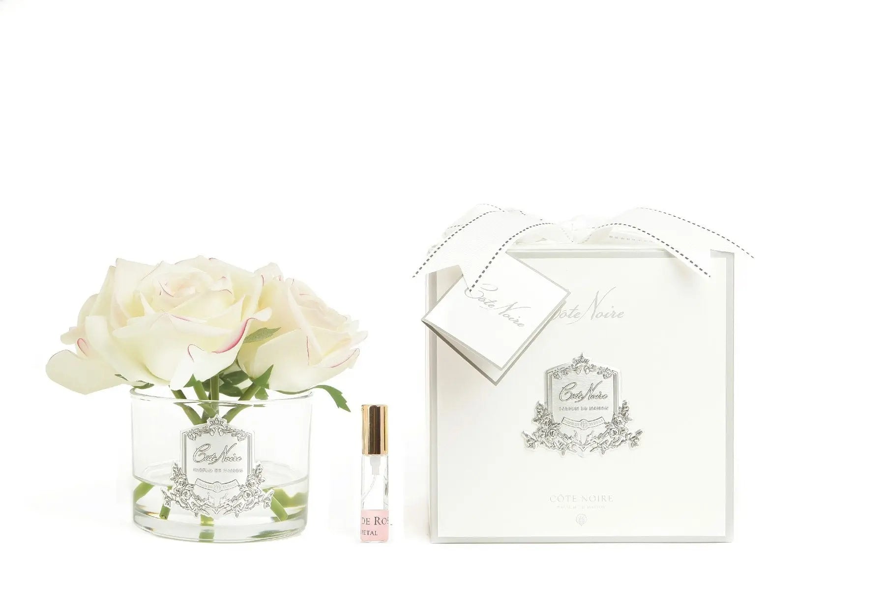 A Cote Noire gift set featuring five artificial pink blush roses in a clear glass vase with a silver crest. Accompanying the vase is a small vial of perfume labeled 'Pétale de Rose' with a gold cap. The items are displayed next to an elegant white gift box adorned with a silver emblem and a white ribbon, creating a refined and luxurious presentation.