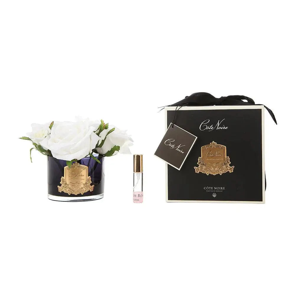 The image showcases an elegant product display including a floral arrangement and a fragrance set. On the left, there is a dark blue glass container embellished with a gold crest, filled with pristine white artificial roses. Next to it, a small clear perfume vial labeled with gold text is presented. On the right, a sophisticated black box with gold detailing and a black ribbon tie holds a matching card with the brand logo. 