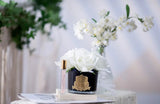The image displays an elegant home decor setting featuring a luxury candle and fragrance product. In the foreground, there is a small, dark blue glass container adorned with a gold crest, holding a large white artificial rose. Beside it, there's a slender perfume vial with a pink label and golden cap. The setting includes a blurred background with delicate white flowers in a vase, enhancing the romantic and sophisticated atmosphere.