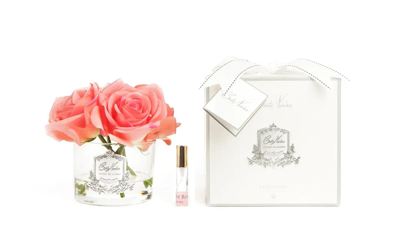 The image features a sophisticated perfume presentation. On the left, there are several vibrant pink roses arranged in a clear glass vase with a label that reads "Cote Noire Bougie de Jardinia." Beside the vase is a small, elegant roll-on perfume bottle with a label matching the vase. On the right, there is a white gift box with a ribbon and a gift tag labeled "Cote Noire." The box and the tag both display ornate, silver decorative elements, highlighting the luxurious nature of the products.