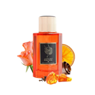 This is an image of a rectangular, transparent orange bottle of perfume labeled "Golden Amber" by Ibraheem Al Qurashi. The perfume bottle has a white cap and features black Arabic calligraphy on the label. Beside the bottle, there's a peach-colored rose, a slice of dried orange, a few cloves, and some pieces of amber resin, all placed against a white background. 