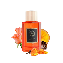 This is an image of a rectangular, transparent orange bottle of perfume labeled "Golden Amber" by Ibraheem Al Qurashi. The perfume bottle has a white cap and features black Arabic calligraphy on the label. Beside the bottle, there's a peach-colored rose, a slice of dried orange, a few cloves, and some pieces of amber resin, all placed against a white background. 