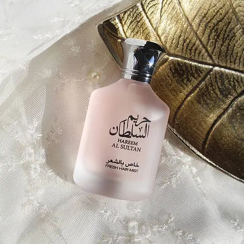 The image features a frosted pink perfume bottle with a metallic cap, placed on a textured surface, possibly silk, with a golden-colored fabric or object partially visible in the background. The bottle is labeled with "HAREEM AL SULTAN" in both English and Arabic script, and below it reads "FRESH HAIR MIST." The setting and the luxurious appearance of the bottle suggest it is a high-end hair fragrance product.