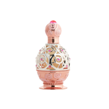The image depicts an ornate perfume bottle on a white background. The bottle features a rose gold base and cap with intricate swirl patterns embossed on the surface. A decorative metal overlay with filigree designs encases the clear central portion of the bottle, adorned with small pink gemstone accents. The cap is topped with a pink crystal-like element, adding to the luxurious appearance of the bottle.