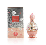 The image shows a perfume bottle next to its packaging. The bottle is identical to the one previously described, with a rose gold and pink design, intricate filigree, and gemstone accents. The packaging box matches the bottle with a rose gold bottom and a greyish top featuring swirling patterns, some with red accents. The box has a label with "Haneen" written in white on a red background and "Concentrated Perfume Oil" below it.