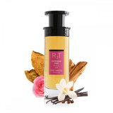 This is an image of a cylindrical clear perfume bottle with a black pump and a yellowish liquid inside, labeled "RT Happiness Dose" with additional text in Arabic and "ROSE TOBACCO" underneath. The bottle is positioned against a white background with a pink rose, a dry tobacco leaf, a cluster of vanilla beans, a white orchid flower, and some cinnamon sticks arranged around it, indicating the scent notes of the perfume.