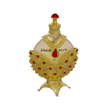 The image displays an opulent perfume bottle with a white background. The body of the bottle is a pale, translucent yellow, topped with a bright red cap resembling a gemstone. The bottle is adorned with a golden overlay featuring intricate cut-out patterns and red gemstone accents. Arabic script is prominently displayed in the center, with smaller text below.
