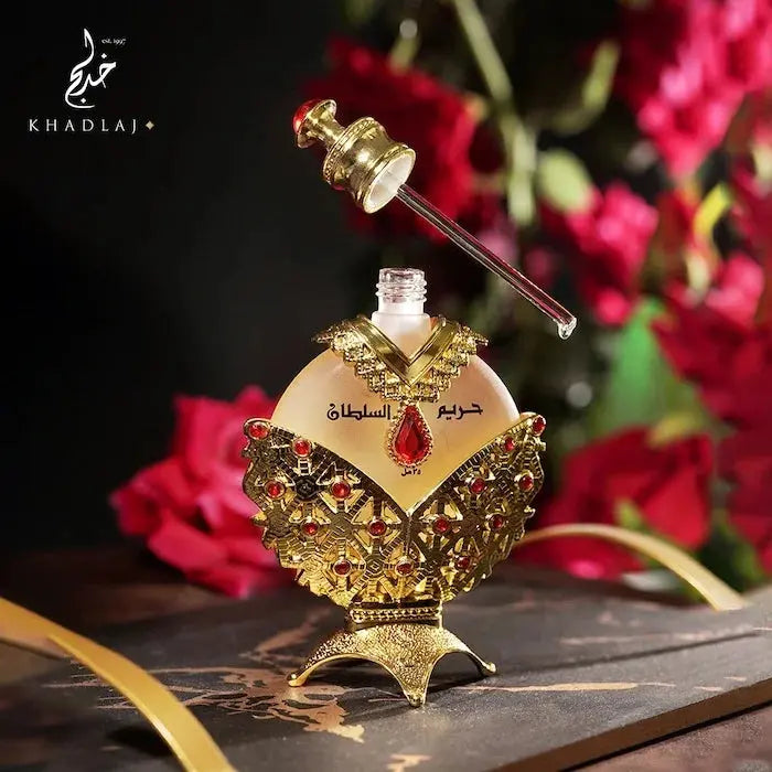 The image presents a luxurious perfume bottle with the cap removed, revealing a glass applicator attached to the golden cap. The bottle has a heart-shaped translucent yellow body, ornately decorated with a golden filigree design embellished with red gemstones. It has an Arabic inscription in the center. The bottle is perched on a textured golden base, designed to give an opulent appearance.