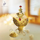 The image shows a beautifully crafted perfume bottle with a cap on, set against a softly lit, bokeh background that creates a warm and luxurious atmosphere. The translucent heart-shaped bottle is adorned with a golden filigree design and red gemstones. The cap is topped with a red gemstone, and the bottle rests on an elaborate golden base. In the foreground, the bottle's intricate golden cap lies detached next to the bottle, mirroring its design.