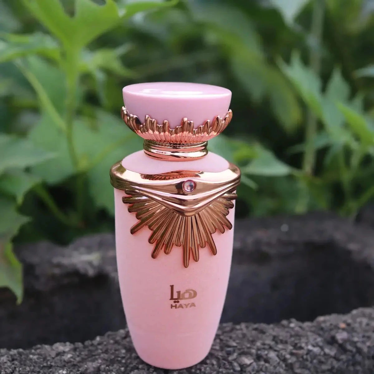 The image depicts a perfume bottle positioned outdoors with green foliage in the background. The bottle has a matte pink body and a rose gold neck and cap. The cap is adorned with a distinctive wing-like design and a small jewel-like detail on the front, hinting at a luxurious style.  The overall setting suggests a natural and fresh ambiance, potentially aligning with the fragrance's character.