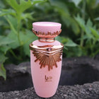 The image depicts a perfume bottle positioned outdoors with green foliage in the background. The bottle has a matte pink body and a rose gold neck and cap. The cap is adorned with a distinctive wing-like design and a small jewel-like detail on the front, hinting at a luxurious style.  The overall setting suggests a natural and fresh ambiance, potentially aligning with the fragrance's character.