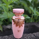 The image depicts a perfume bottle positioned outdoors with green foliage in the background. The bottle has a matte pink body and a rose gold neck and cap. The cap is adorned with a distinctive wing-like design and a small jewel-like detail on the front, hinting at a luxurious style.  The overall setting suggests a natural and fresh ambiance, potentially aligning with the fragrance's character.