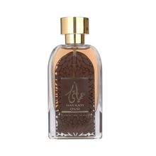 The image shows a clear glass perfume bottle with a golden cap. The perfume liquid is a dark amber color. The front of the bottle features a decorative motif resembling a dome with geometric patterns in brown and a central label with the name "HAYAATI OUD" in gold lettering and elegant Arabic calligraphy. The overall design suggests an opulent and exotic fragrance, and the bottle is set against a plain white background to emphasize its design and contents.