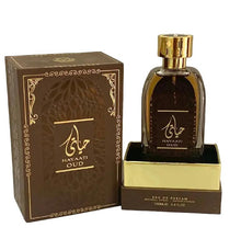 n image of Hayaati Oud perfume by Ard Al Zaafaran, featuring the perfume bottle and packaging. The bottle is transparent with a dark amber liquid and a reflective gold cap. It sits on a gold-colored base with the text "EAU DE PARFUM NATURAL SPRAY 100ml e 3.4fl.oz" at the bottom. The brown box has an ornate design with golden accents and Arabic calligraphy, along with the name "HAYAATI OUD" in both Arabic and English.