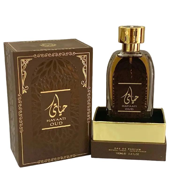 n image of Hayaati Oud perfume by Ard Al Zaafaran, featuring the perfume bottle and packaging. The bottle is transparent with a dark amber liquid and a reflective gold cap. It sits on a gold-colored base with the text "EAU DE PARFUM NATURAL SPRAY 100ml e 3.4fl.oz" at the bottom. The brown box has an ornate design with golden accents and Arabic calligraphy, along with the name "HAYAATI OUD" in both Arabic and English.